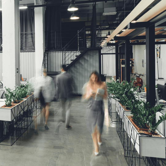 People walking through a modern, open-plan office space with plants, desks, and a stairway in the background. The image is slightly blurred, indicating movement.